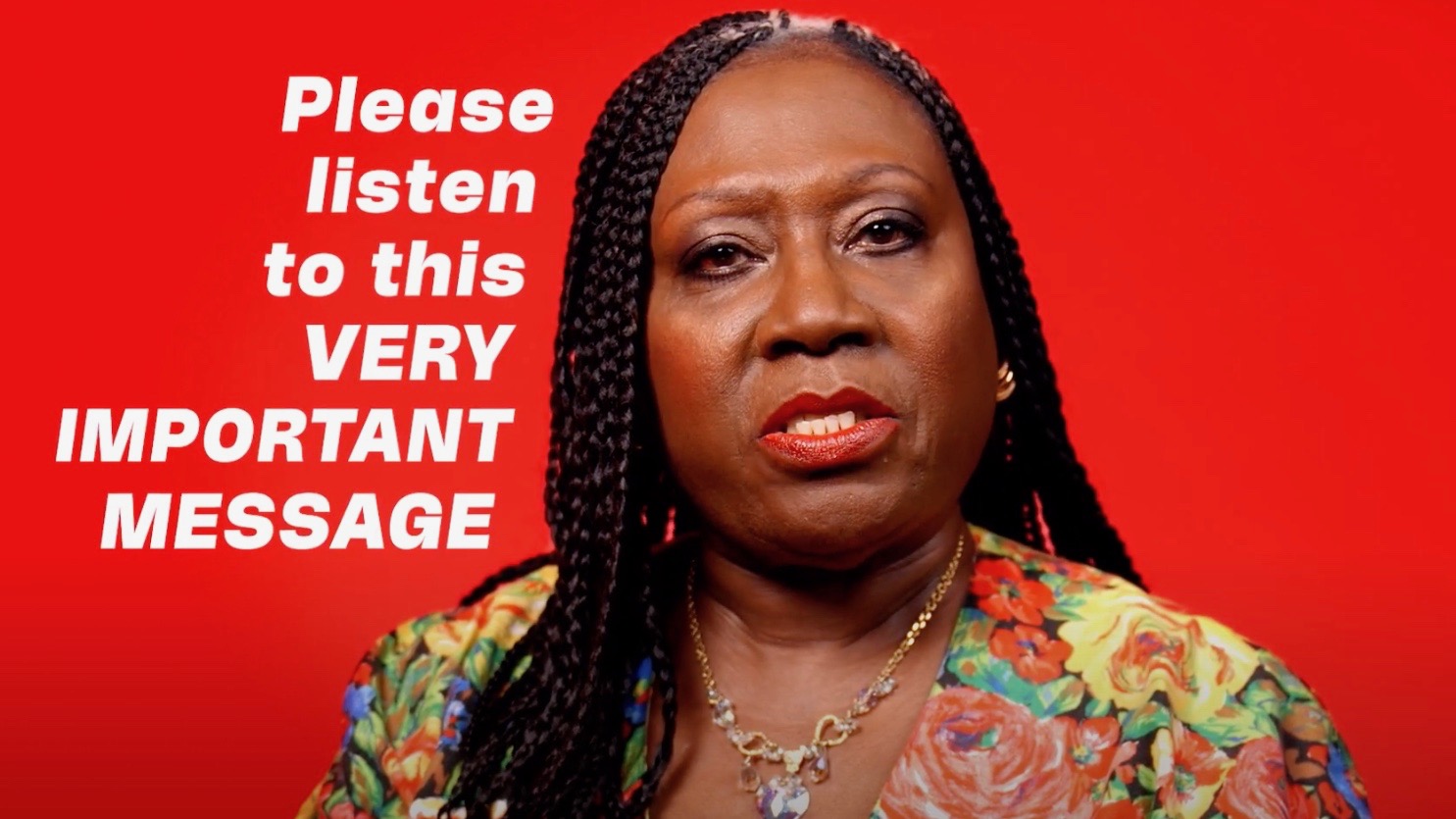 A black lady is facing forwards. Against a bright red background, text in white says 'Please listen to this very important message'