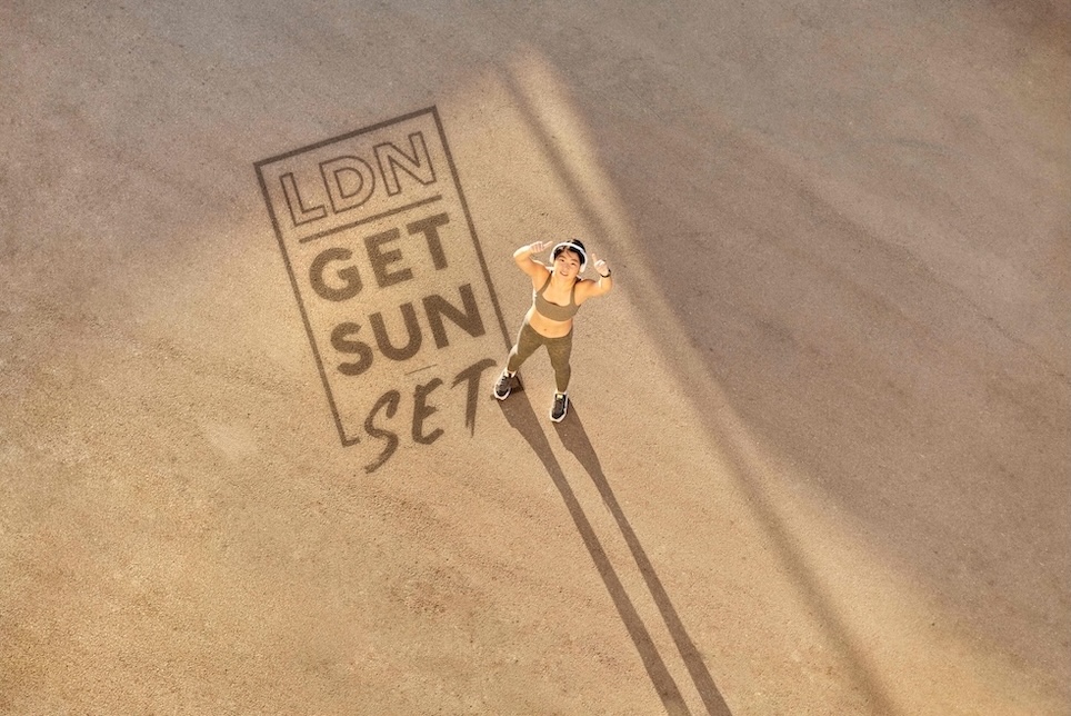 A runner is on sand staring up at the sky and giving the thumbs up. On the sand is printed the words LDN get sun set.