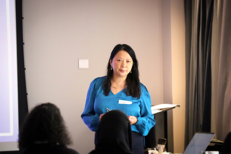 A woman in a blue top is standing facing an audience who are seated.