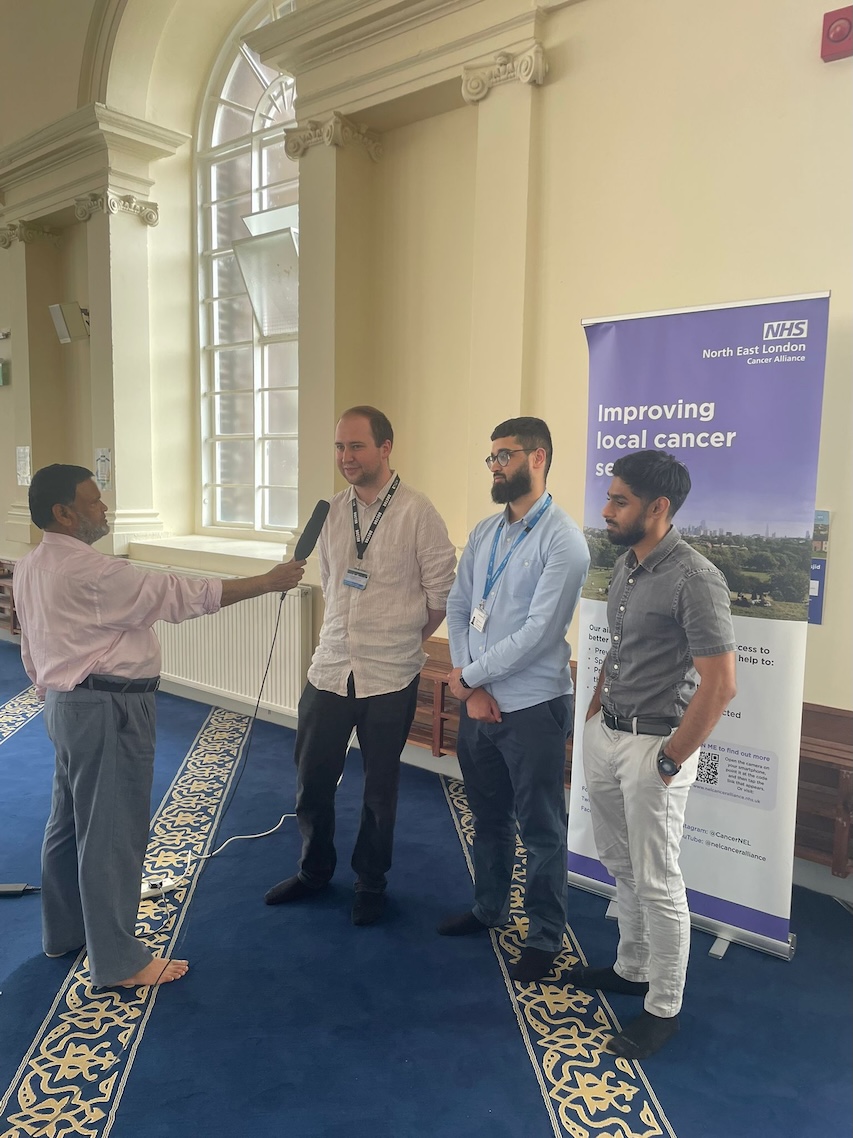 Three men are being interviewed by a man with a microphone. They are standing in front of a banner which says Improving Local Cancer Services.