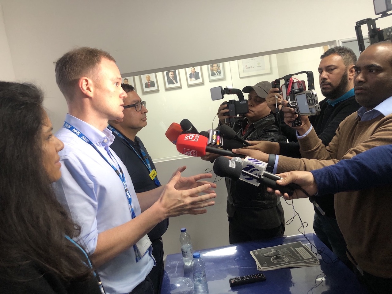 A panel of speakers is standing in front of people with cameras and microphones.