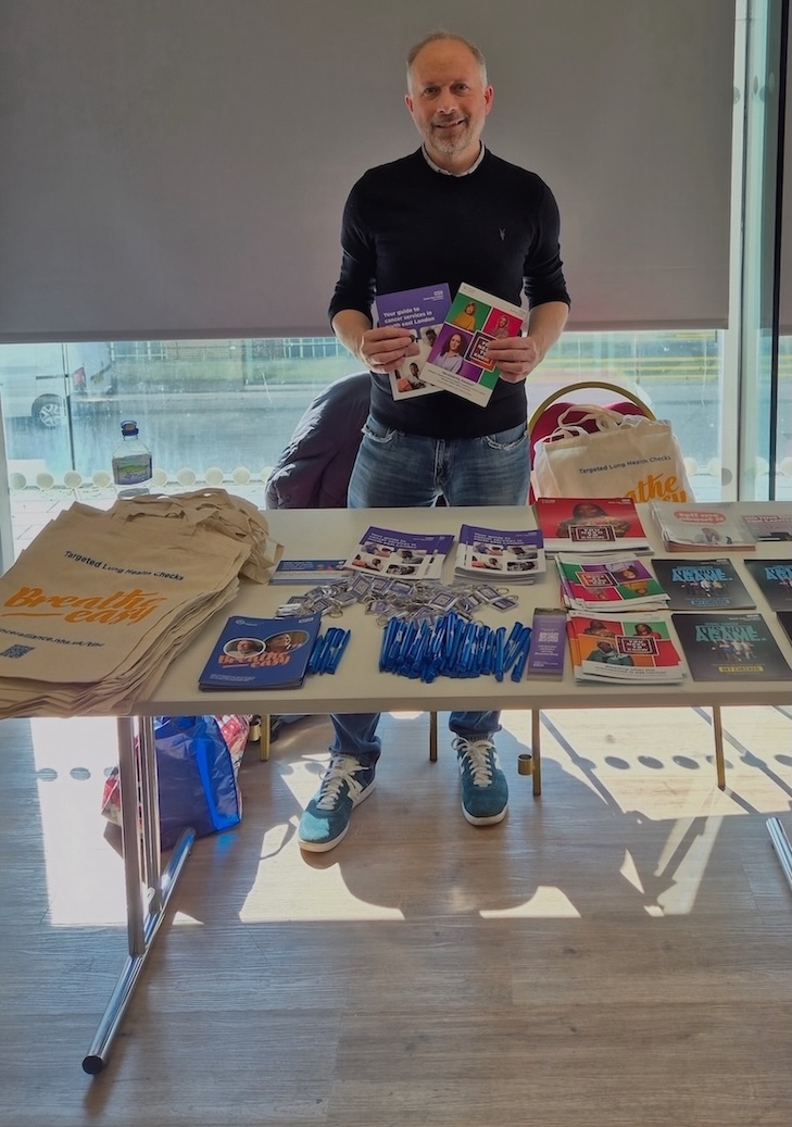 A man is standing in front of a table with a range of cancer information leaflets and is holding a couple of leaflets up to the camera