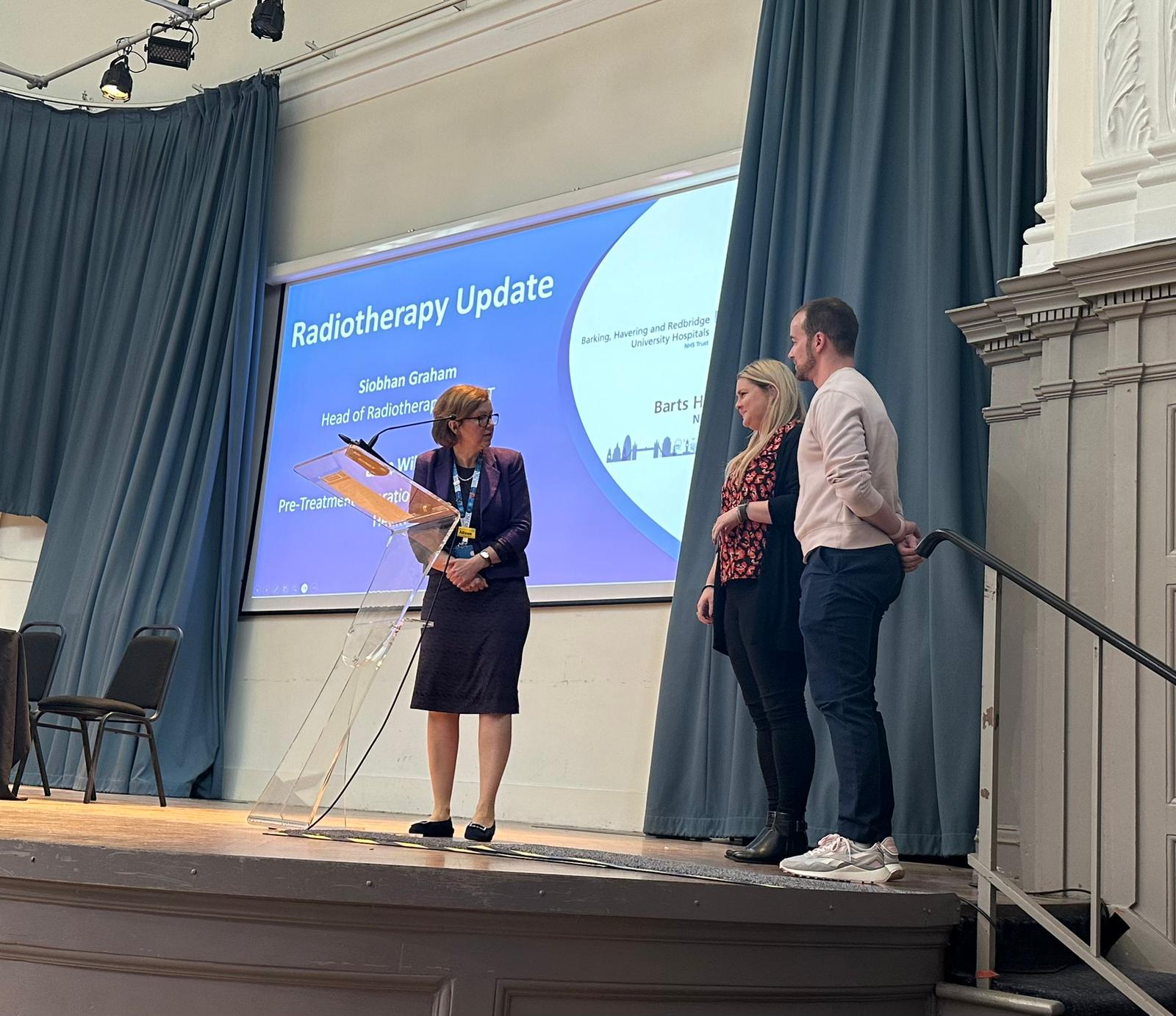 A lady is on a stage introducing two guests who are going to give a presentation to an audience.