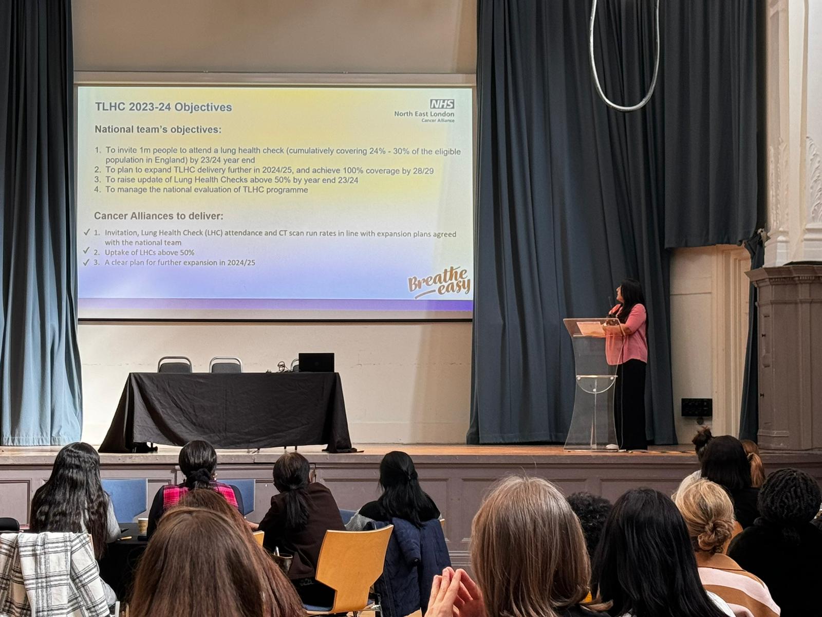 A lady is on a stage giving a presentation to an audience