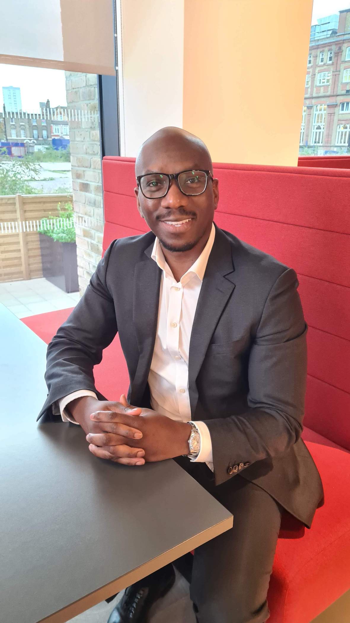 A black male is sat at a table wearing glasses and a suit