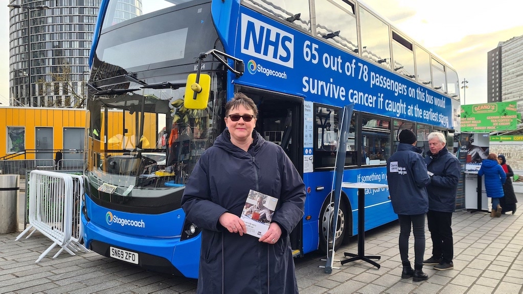 A lady is standing in front of a blue double-decker NHS bus