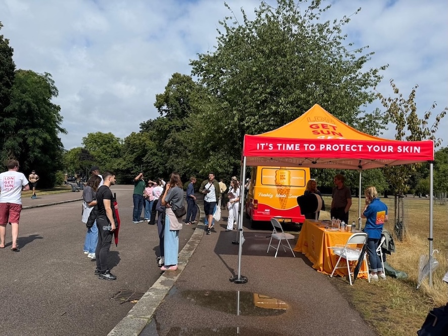 A gazebo is in a park with the words It's Time To Protect Your Skin on it and crowds of people are gathered near it.