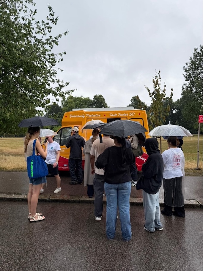 A crowd is queuing in the rain holding umbrellas and waiting for free sun cream from a yellow ice cream van.