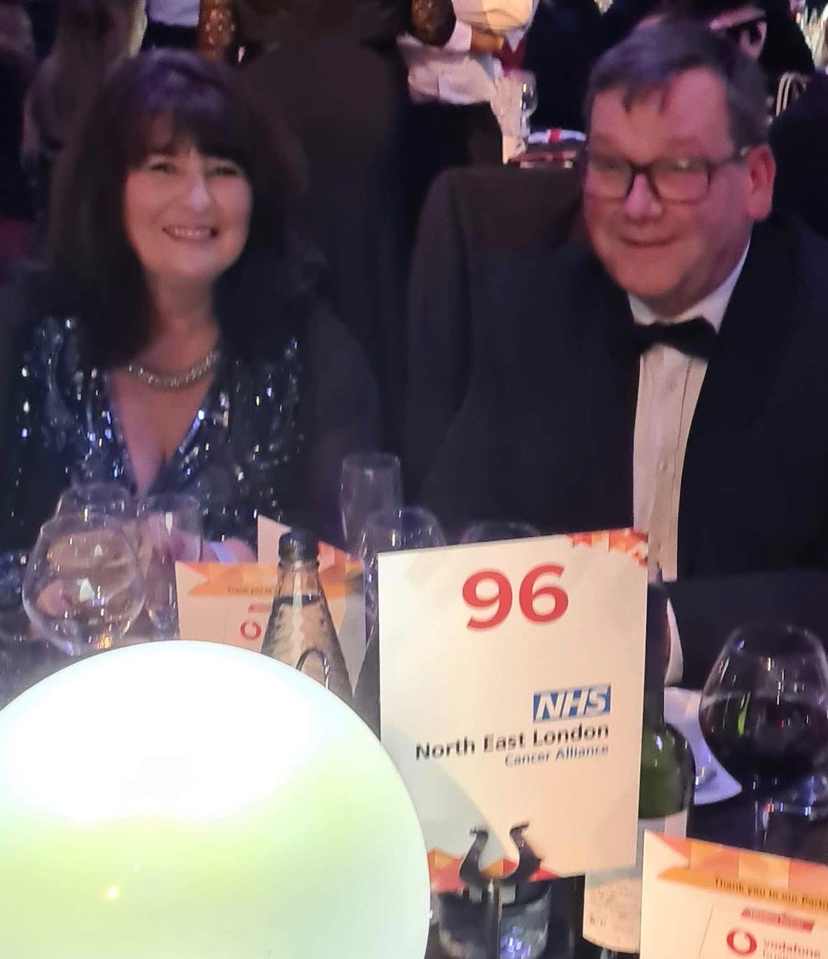 A man and a woman are dressed smartly and are sitting at an awards event dinner table. On the table is a sign which has the number 96 on it and the words North East London Cancer Alliance