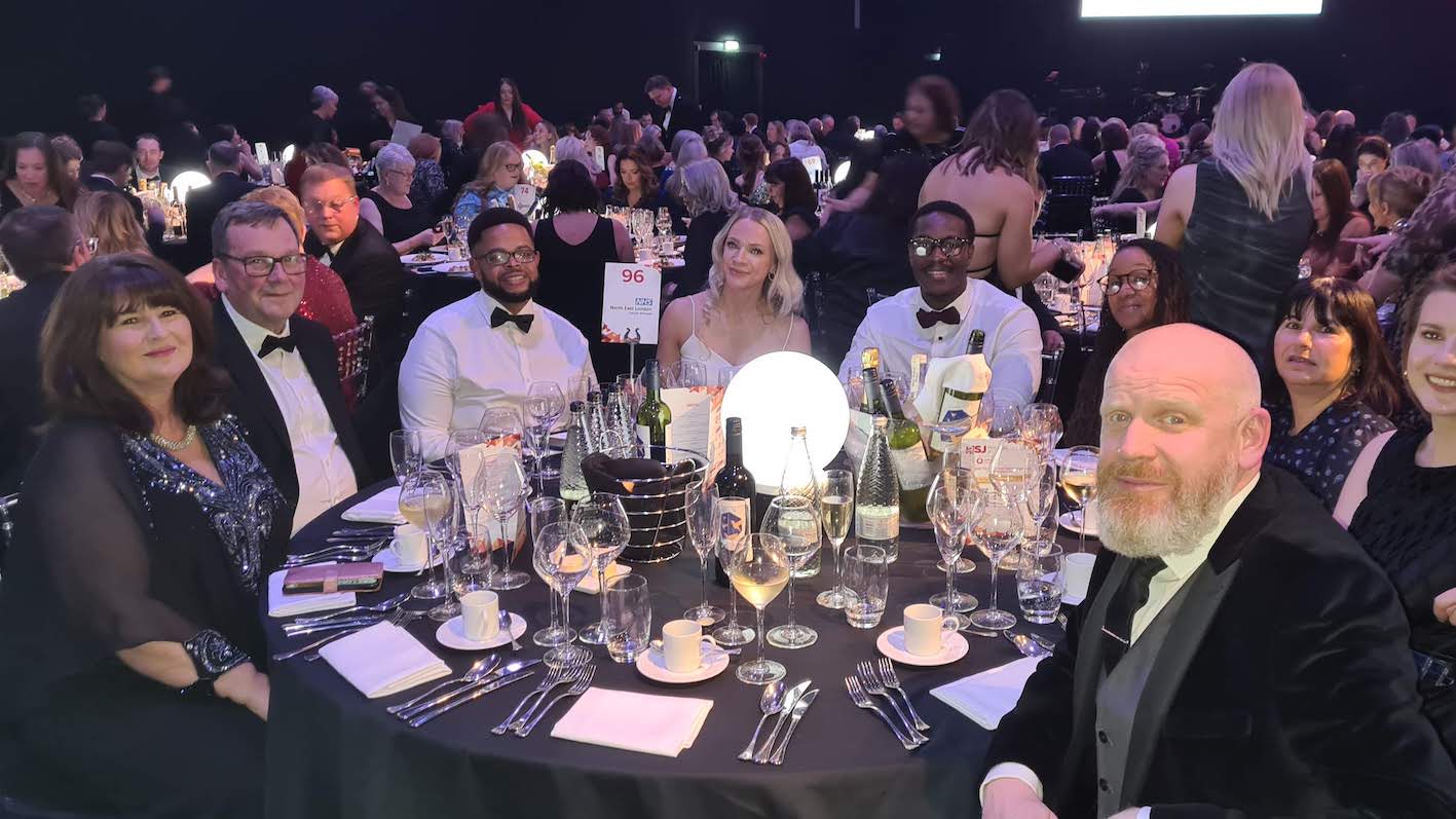 A group of 9 people are sat at an awards event dinner table dressed smartly and facing the camera.