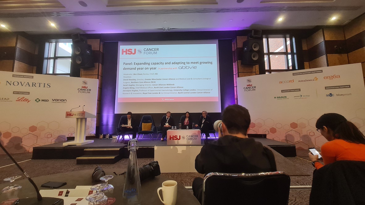 Five people are sat on a stage as a panel of speakers. Behind them is a big screen which says HSJ Cancer Forum on it. In the foreground you can see a photographer taking a photo of the panel.