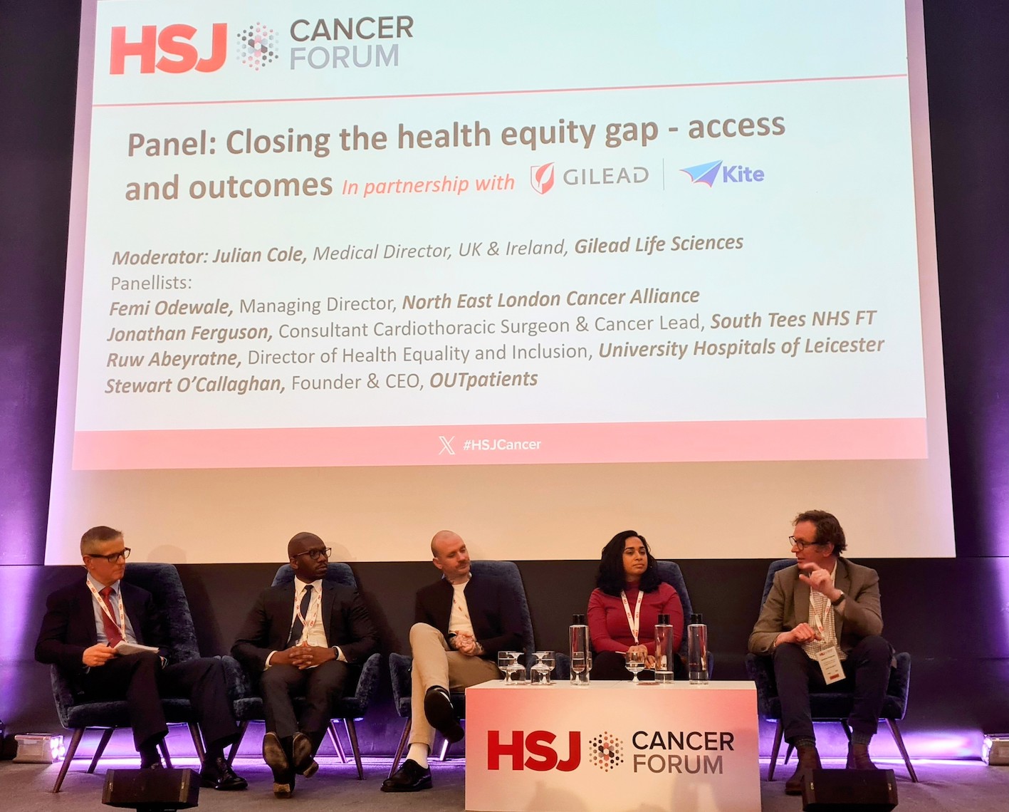 Five people are sat on a stage as a panel of guest speakers with a large screen behind them which says HSJ Cancer Forum on it