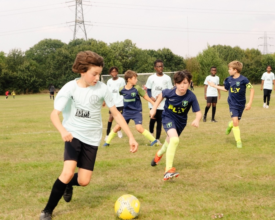 The image shows a football match taking place between two team of boys. One team is wearing white shirts with It's Not a Game written on them, The other team is wearing blue kit.