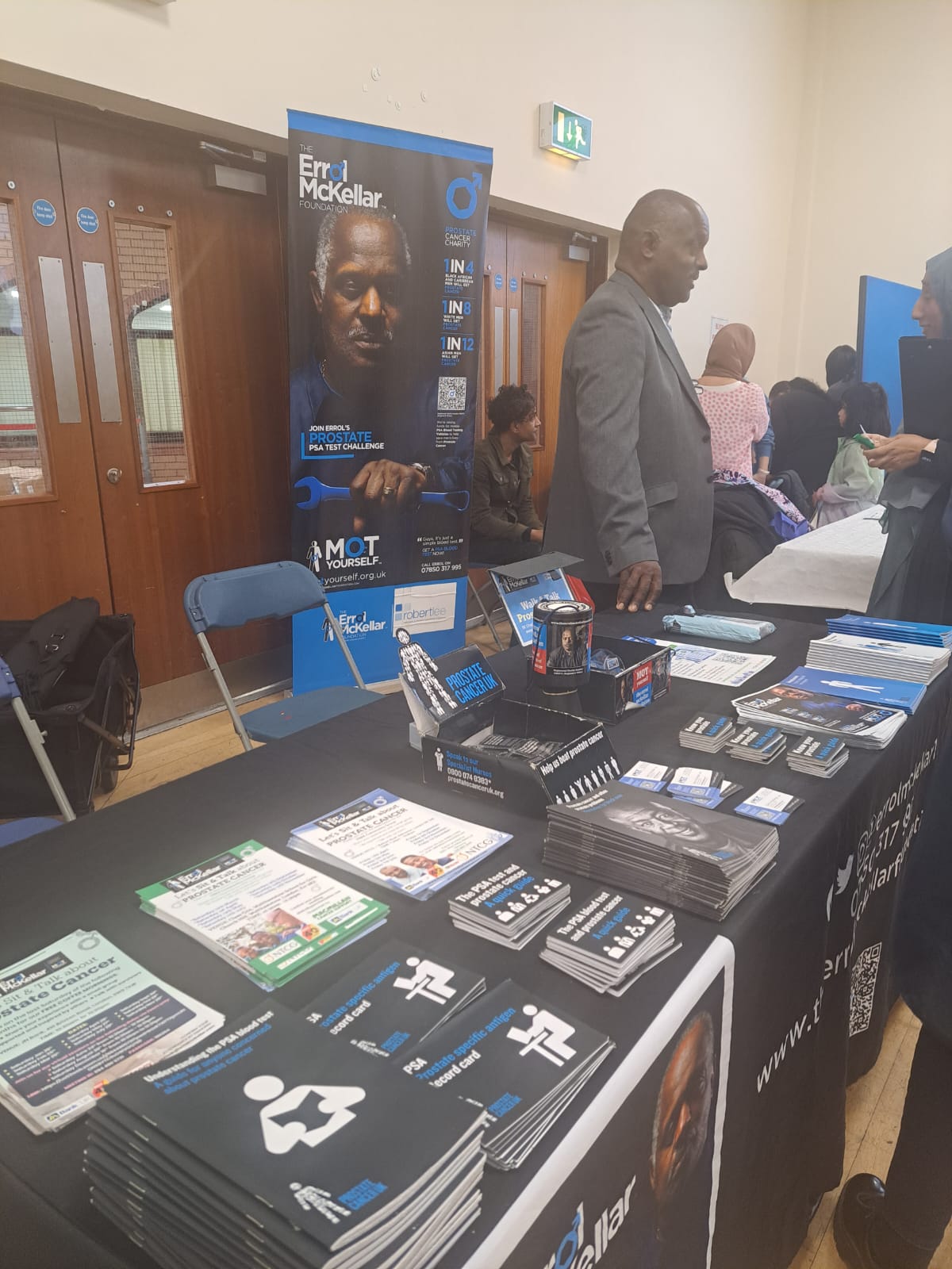 A black man is talking to people about prostate cancer at an event. He is standing by a table with leaflets on it. A banner behind him says The Eroll McKellar Foundation