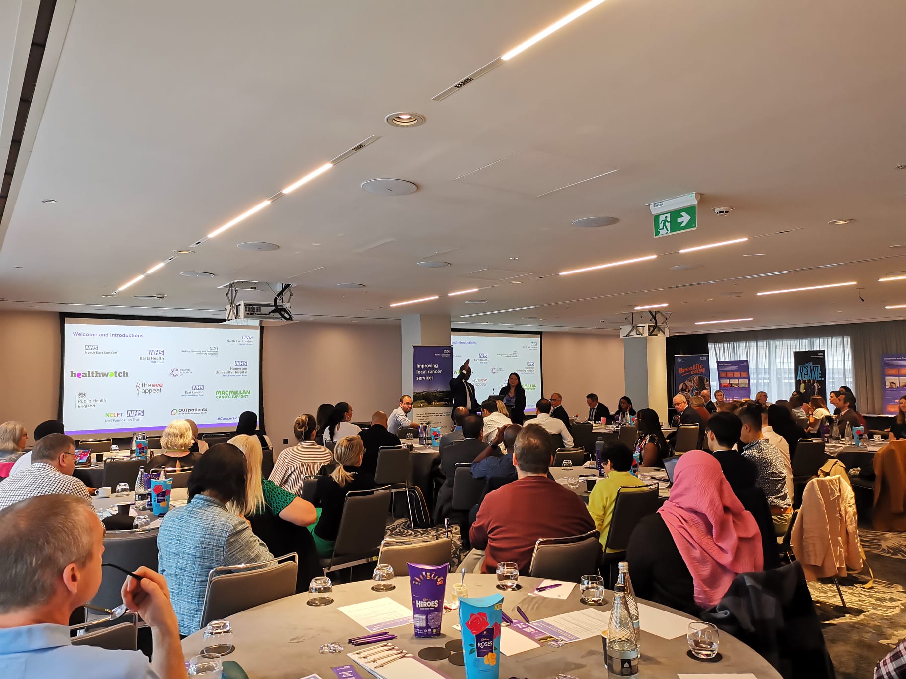 A man and a woman are standing at the front of a large conference centre giving a presentation. The audience are seated at round tables.
