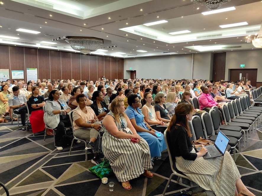 A conference hall packed with people sat in chairs facing forwards.