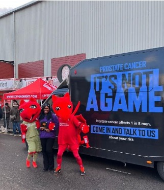 A person with two mascots is standing next to a van which says It's Not A Game on it