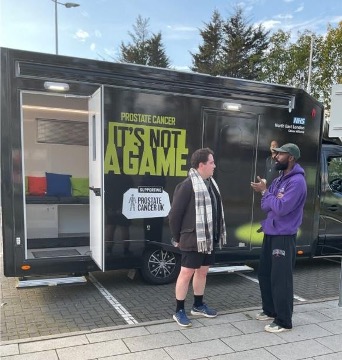 Two men taking in front of a van which has It's Not A Game on it