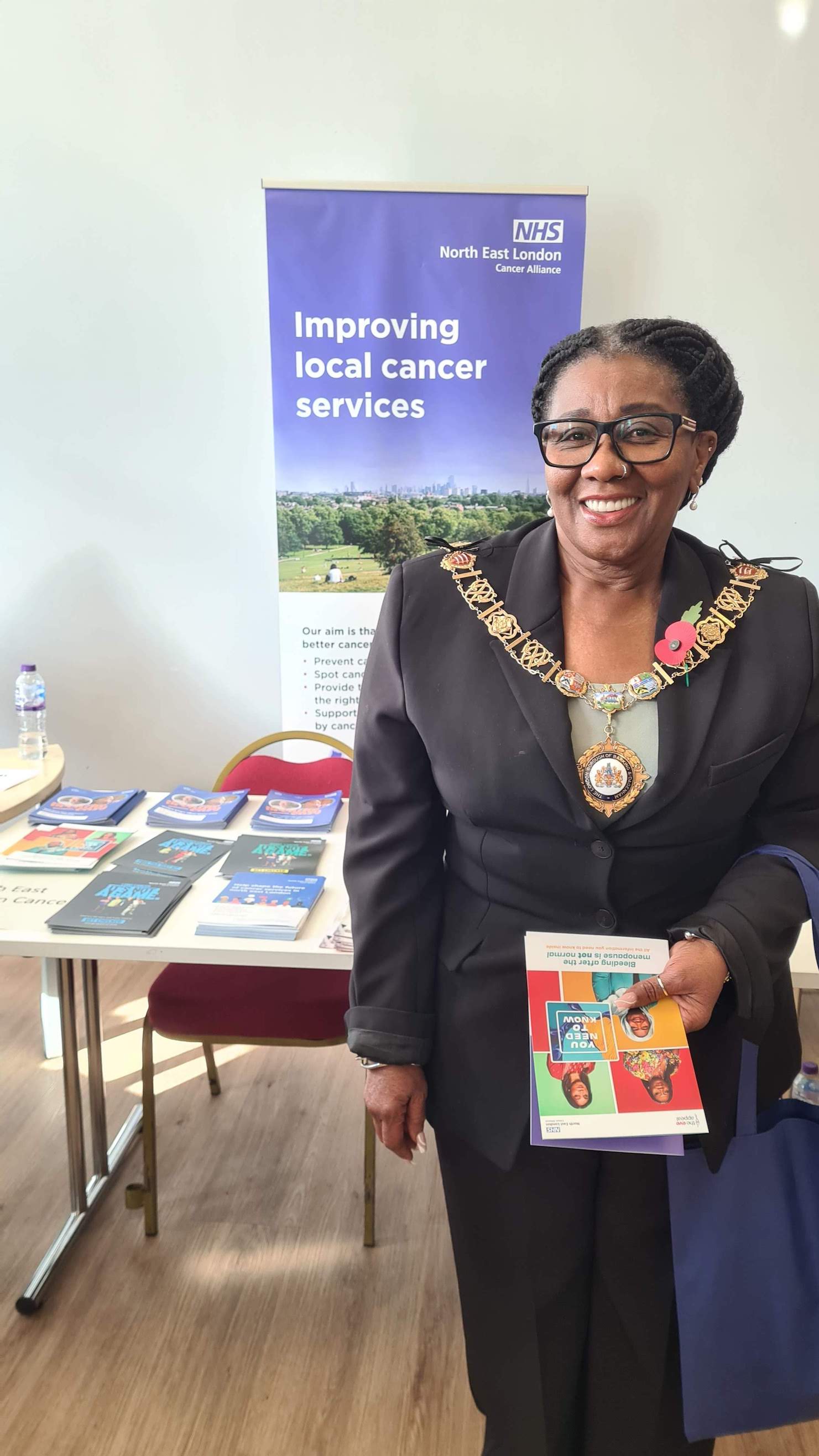 The mayor of Barking Council is standing in front of an exhibition stand which says Improving Local Cancer Services on it. She is holding a couple of cancer information leaflets.
