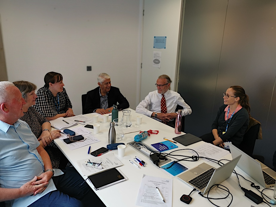 A mixed group of six people are sat around a table in a business meeting having a discussion with laptops on the table in front of them.