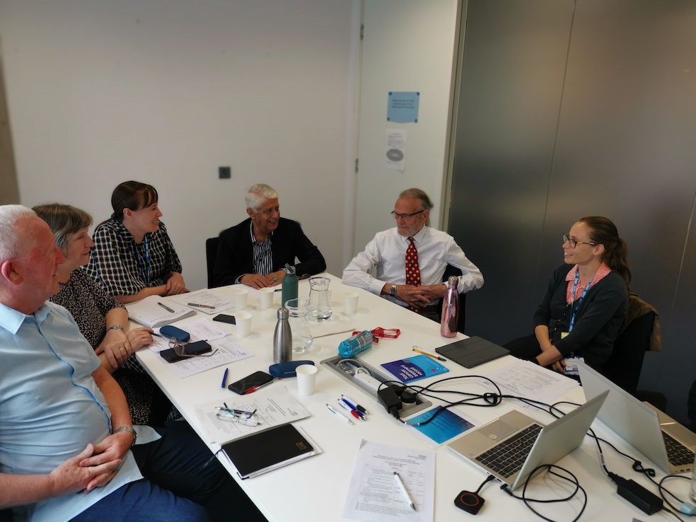 A group of patient and carer representatives are sat around a table having a meeting. On the table are laptops and notepaper.