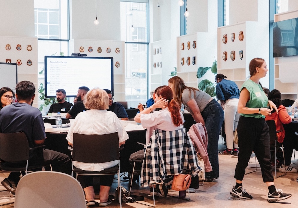 A large room with people having discussions around tables with a presentation screen in the background.