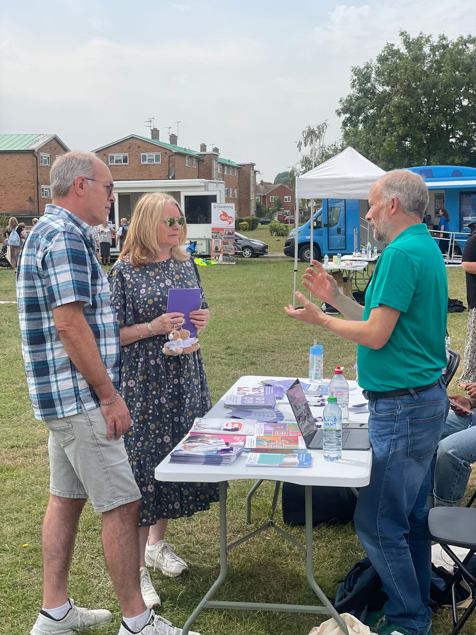 A man is talking to two people outside at an event behind a table with information leaflets on it