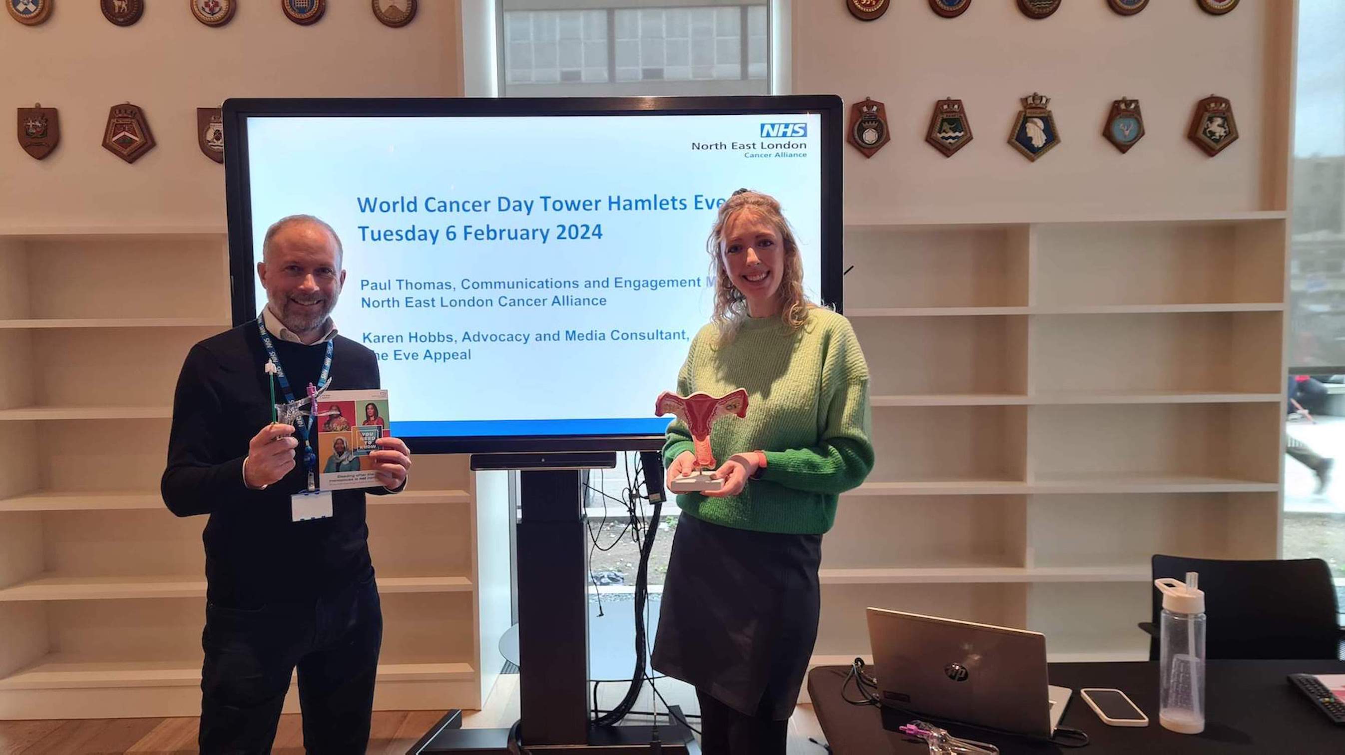 A man and a lady are standing in front of a large TV screen. On the TV screen is a PowerPoint slide which says World Cancer Day Tower Hamlets Event on it. The man and lady are holding information and materials about womb cancer and cervical screening