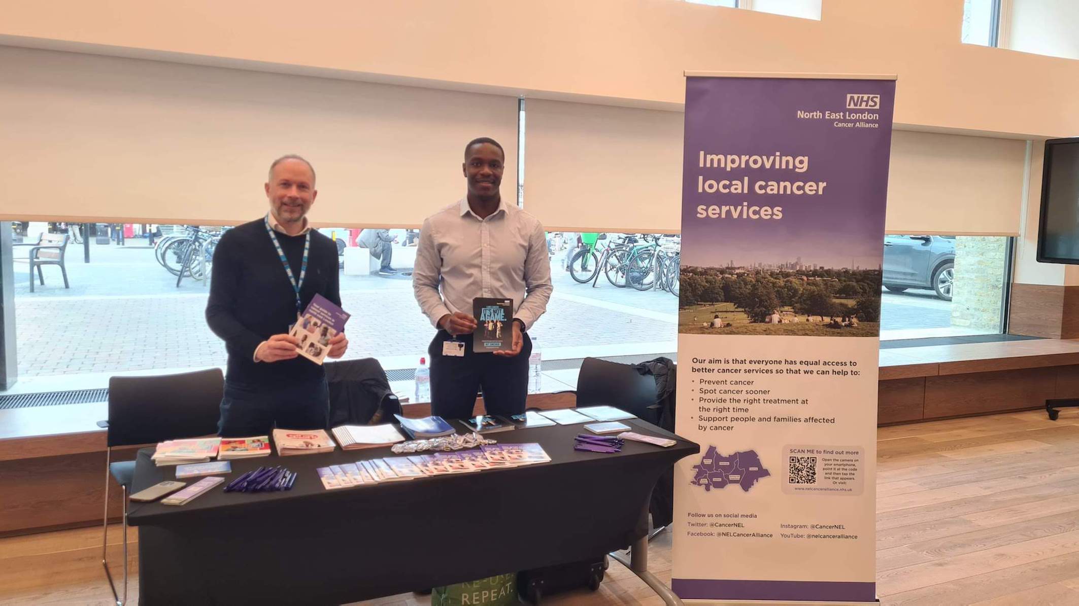 Two men are standing in front of a cancer awareness stand which has patient leaflets on it.  A pop up banner says 'Improving Local Cancer Services' on it.