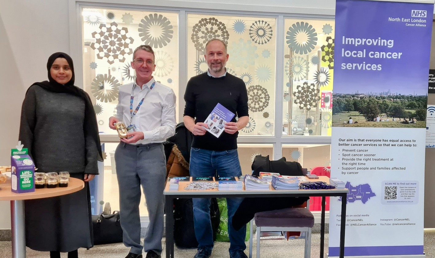 Two men and a lady are standing at cancer information stand which has cancer leaflets displayed on a table. A pop-up banner has the words 'Improving local cancer services' on it.