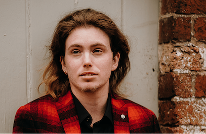 A person is standing against a white door near a brick wall, facing the camera. He is wearing a red checked shirt