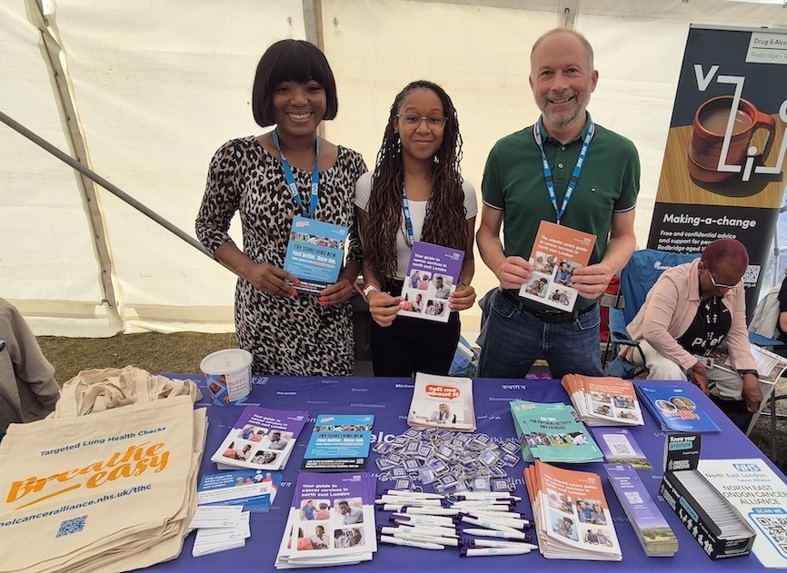 Three people are standing behind a table which has cancer information leaflets on it.