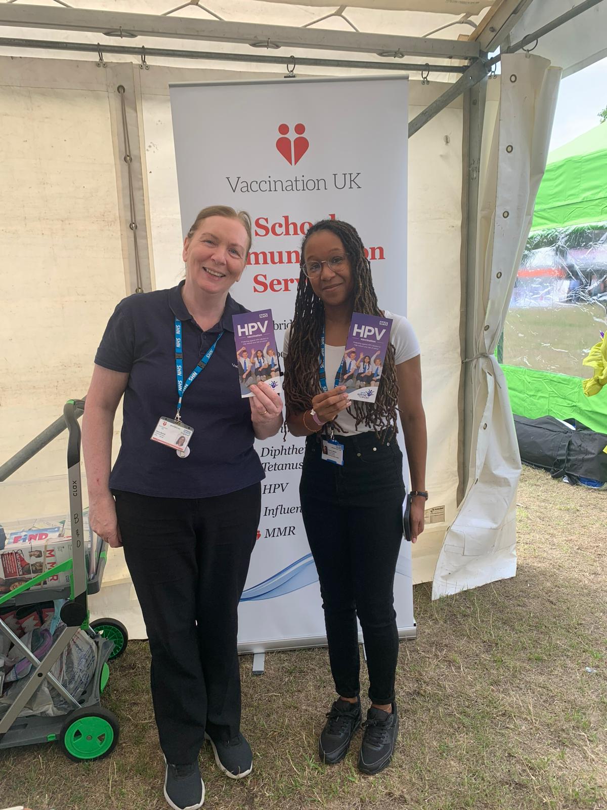 Two women are standing holding a leaflet about HPV in front of a banner which says Vaccination UK.