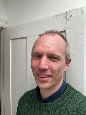 A man wearing a green jumper is standing in front of a white door.