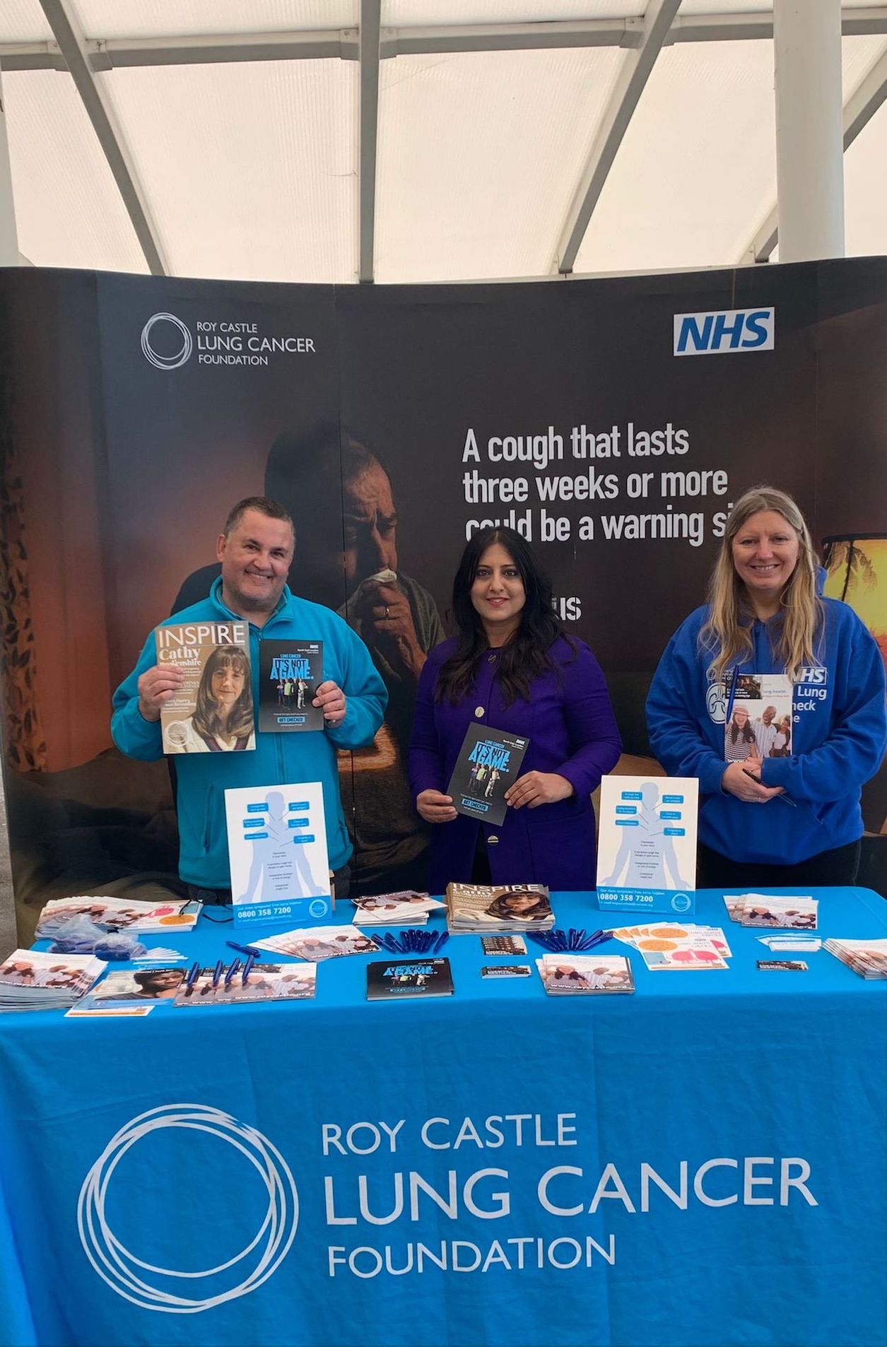 Image of three people standing behind a desk holding up leaflets which have cancer information on them. The banner on the table has the words "Roy Castle Lung Cancer Foundation' on it.