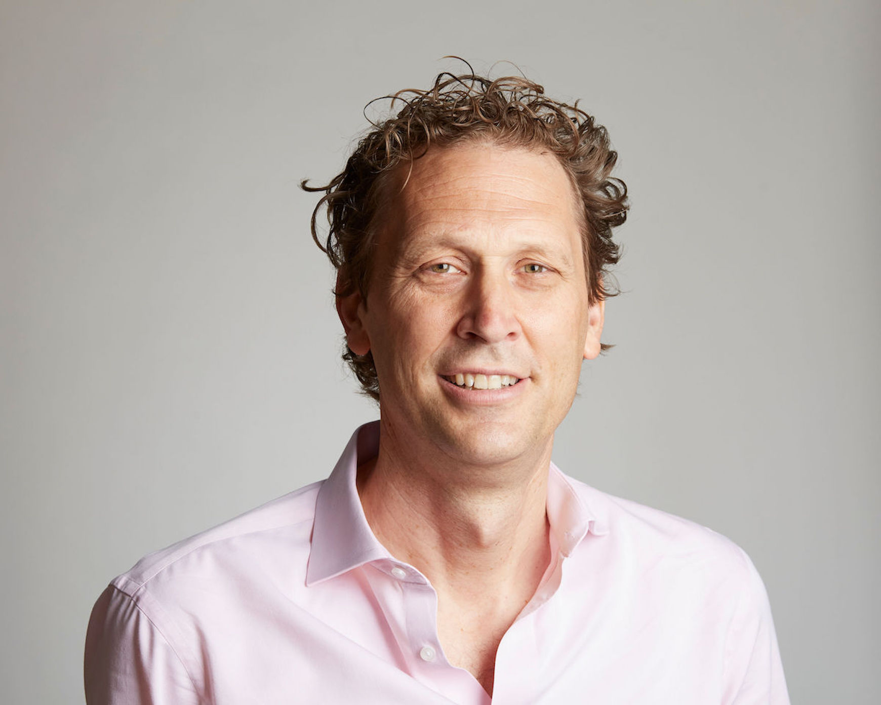 The image is of a middle-aged white male wearing a smart pink shirt looking towards the camera.