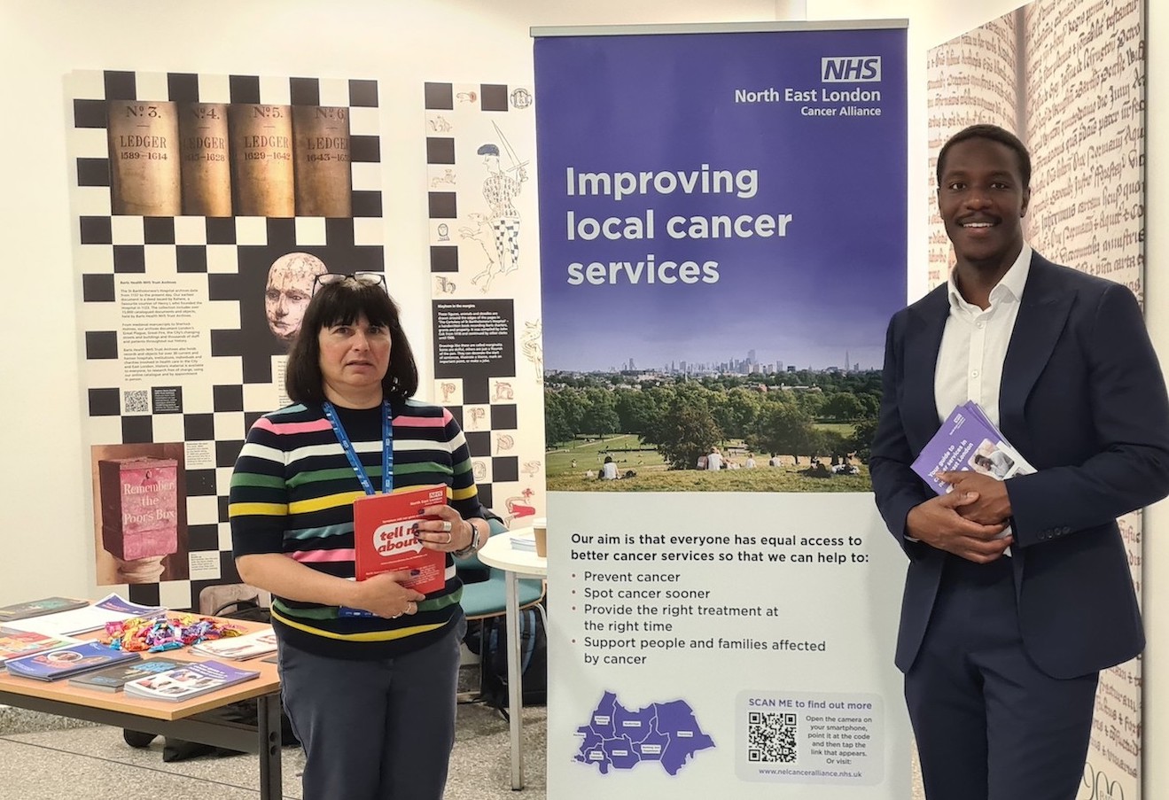 Two people are standing in front of a table with cancer information leaflets on it. To the right is a banner which says improving local cancer services.