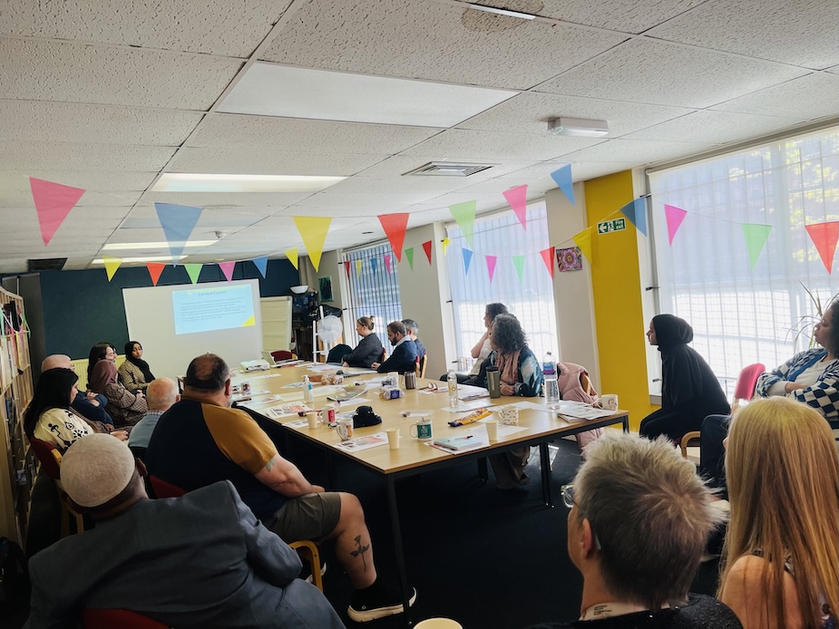 A number of people are sat around a table in a large meeting room looking at a presentation screen which is at the far end of the room.