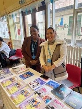 Two women are standing behind a table which has cancer information leaflets on it.