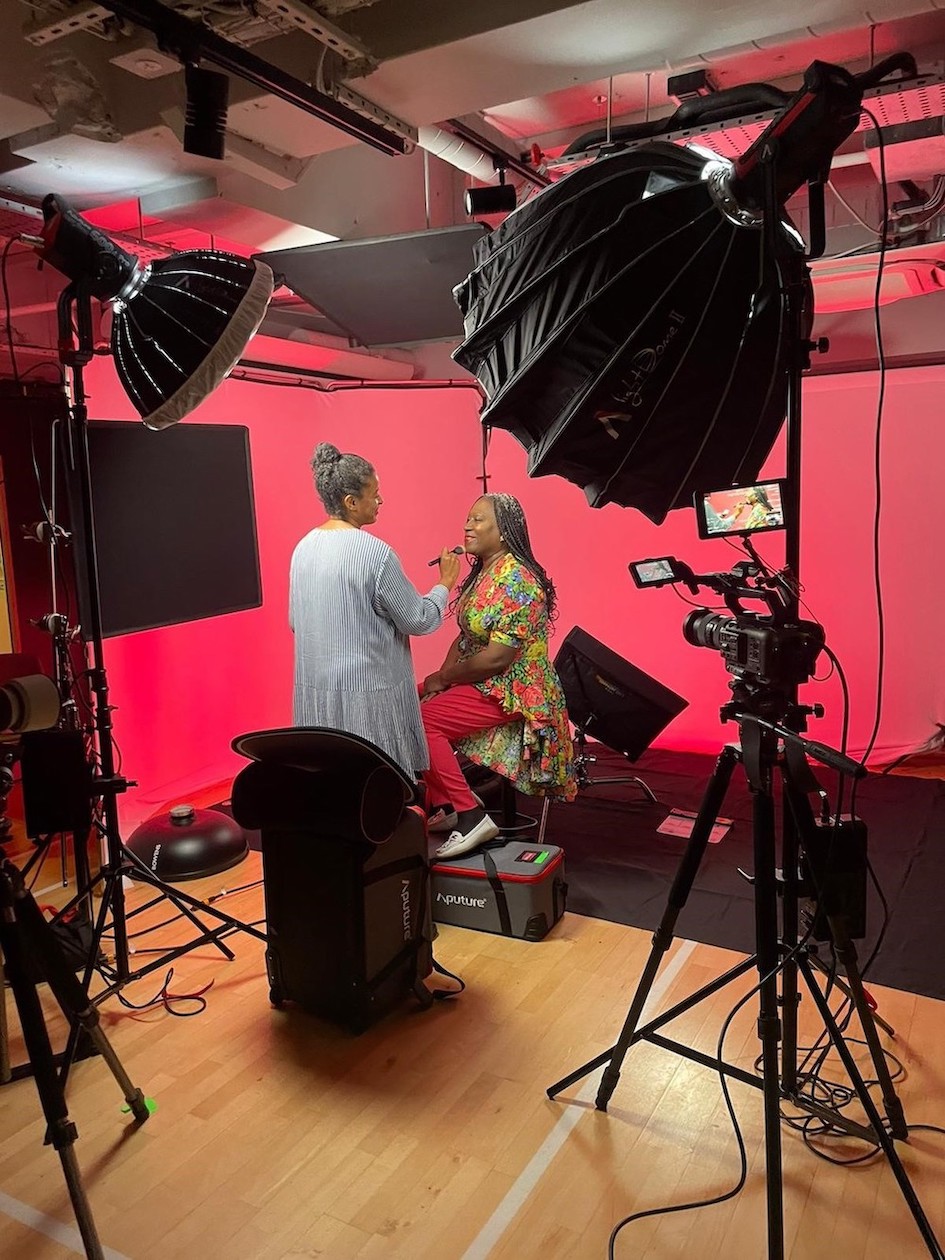 A lady is sat on a stool in what looks like a film studio with a camera and lights pointing at her. Another woman is standing in front of her, helping her do her make up, getting her ready for the photo shoot. 