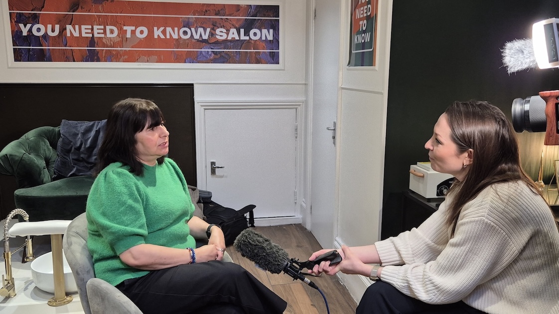 A lady in a green top is being interviewed by a lady holding a microphone. They are both sat in a nail salon