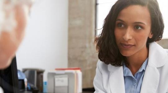 A lady sat at a desk facing forward. She is talking to what looks like an older lady with white hair.