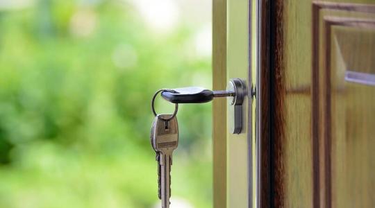A key is shown hanging out of a lock on a front door of a house. A garden is in the background.