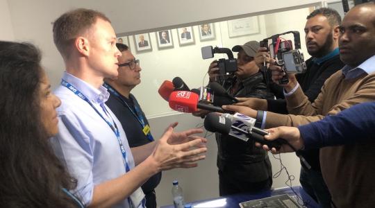 A panel of speakers is standing in front of people with microphones and cameras.