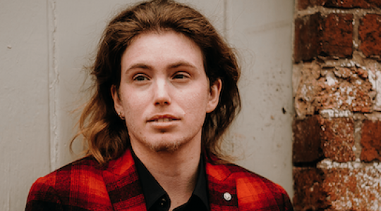 A person wearing a red checked shirt is standing in front of a white door with bricks on the right.