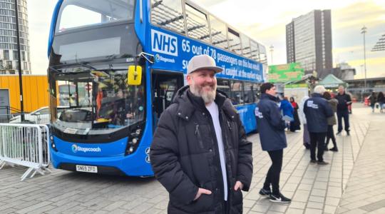 A man is standing in front of a blue NHS double decker bus