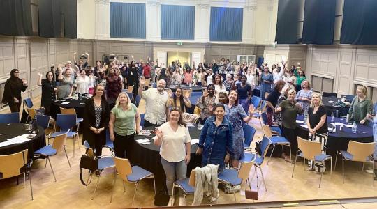 A large number of people are standing in a hall around tables at an event looking up at the camera and waving.