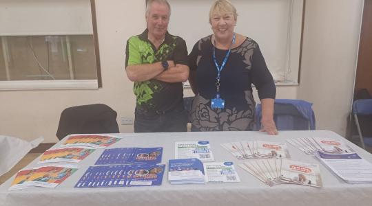 Two people are standing behind a table which has cancer awareness leaflets on it.