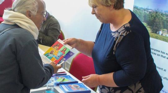 A lady is standing behind a table which has cancer information leaflets on it. She is handing a leaflet to an elderly asian woman and they are having a conversation.