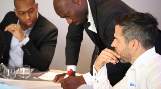 A man in a black suit is standing at a table holding a red marker pen and writing on a flip chart paper. Two other men are sat at the table,  one on either side of him.